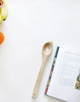 A kitchen scene featuring a wooden spoon placed beside an open cookbook with a recipe for avocado salad with fresh herbs and pepitas. On the left, there's a bunch of bananas, a green apple, and an orange arranged on a clean, white surface.