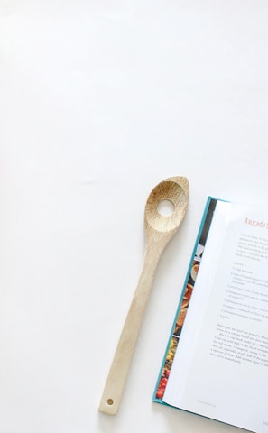 A kitchen scene featuring a wooden spoon placed beside an open cookbook with a recipe for avocado salad with fresh herbs and pepitas. On the left, there's a bunch of bananas, a green apple, and an orange arranged on a clean, white surface.