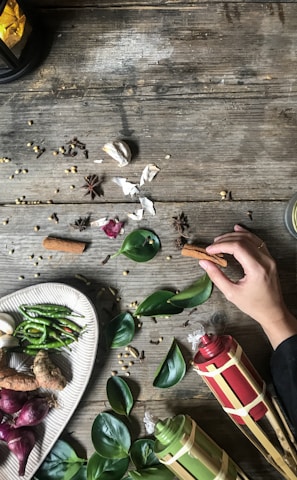 A rustic wooden table is adorned with various culinary ingredients and decorative elements. A hand reaches in to hold a cinnamon stick, surrounded by spices like star anise, garlic, and peppercorns. A plate displays a selection of fresh ingredients including green chilies, shallots, and ginger. Beside it, tropical leaves and traditional decorative items, possibly torches or lanterns, enhance the aesthetic. The setting suggests a theme of natural and traditional cooking or celebration.