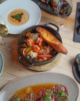 A variety of gourmet dishes are artistically arranged on a wooden table. A black cast iron skillet contains seafood including shrimp and mussels accompanied by a slice of toasted bread. Nearby, a white plate holds a creamy dish adorned with a small garnish. A rectangular black platter features meat or poultry pieces with sesame seeds and greens. At the bottom of the image, a stoneware dish presents sliced meat served in a flavorful sauce with garnishes of greens and red chili peppers. Two glasses of wine, one white and one red, complement the dining setup.