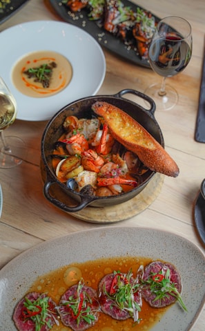 A variety of gourmet dishes are artistically arranged on a wooden table. A black cast iron skillet contains seafood including shrimp and mussels accompanied by a slice of toasted bread. Nearby, a white plate holds a creamy dish adorned with a small garnish. A rectangular black platter features meat or poultry pieces with sesame seeds and greens. At the bottom of the image, a stoneware dish presents sliced meat served in a flavorful sauce with garnishes of greens and red chili peppers. Two glasses of wine, one white and one red, complement the dining setup.