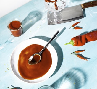 A captivating culinary scene featuring a vibrant setup on a light blue surface. At the center, a white plate holds a glossy brown sauce with a spoon resting on it. Nearby, a large red chili pepper and several smaller peppers are scattered, adding a touch of brightness. A glass jar filled with more of the brown sauce sits to one side, while an empty glass and a cleaver with a wooden handle lie nearby. A crystal glass contains more peppers, creating dynamic shadows across the surface.