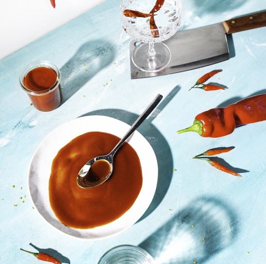A captivating culinary scene featuring a vibrant setup on a light blue surface. At the center, a white plate holds a glossy brown sauce with a spoon resting on it. Nearby, a large red chili pepper and several smaller peppers are scattered, adding a touch of brightness. A glass jar filled with more of the brown sauce sits to one side, while an empty glass and a cleaver with a wooden handle lie nearby. A crystal glass contains more peppers, creating dynamic shadows across the surface.