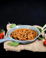A rustic presentation of a dish featuring spiral-shaped cooked ingredients in a blue-gray pan. Accompanying the dish are fresh vegetables including a bunch of lemongrass, a whole garlic bulb, a red tomato, and a small bowl of red and green chili peppers, all arranged on a piece of burlap fabric against a dark background.
