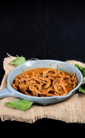 A rustic presentation of a dish featuring spiral-shaped cooked ingredients in a blue-gray pan. Accompanying the dish are fresh vegetables including a bunch of lemongrass, a whole garlic bulb, a red tomato, and a small bowl of red and green chili peppers, all arranged on a piece of burlap fabric against a dark background.