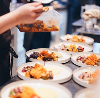 A person is plating food in a kitchen, preparing several dishes with vibrant ingredients. Various plates are arranged on a countertop, each containing a variety of colorful foods, such as vegetables and garnishes.