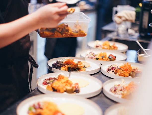 A person is plating food in a kitchen, preparing several dishes with vibrant ingredients. Various plates are arranged on a countertop, each containing a variety of colorful foods, such as vegetables and garnishes.
